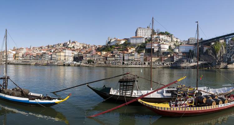 Barcos de colores a lo largo de la orilla del río con una ciudad histórica al fondo.