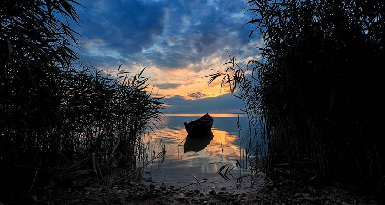 Solitary boat at sunrise framed by reeds with colorful sky reflecting on calm water.