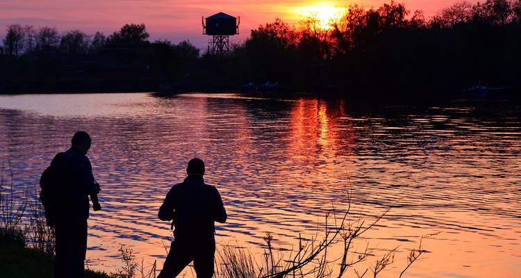 Silhouetted anglers on a riverbank with vivid orange sunset reflecting on the water.