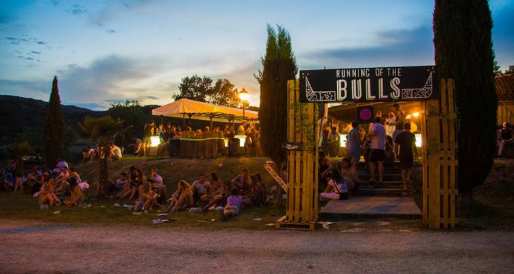 Crowd sitting and standing under a 'Running of the Bulls' sign at sunset.