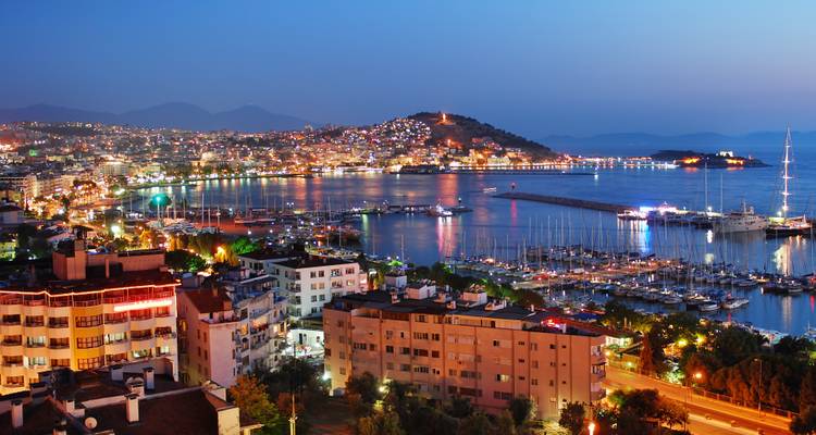 Nachtpanorama des Kusadasi-Yachthafens mit funkelnden Stadtlichtern, die sich auf dem ruhigen Wasser spiegeln.