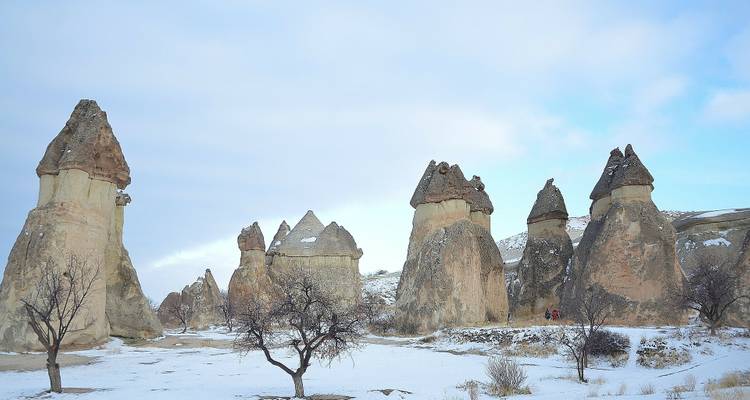 Las chimeneas de hadas cubiertas de nieve se elevan sobre una llanura invernal con dos pequeñas figuras caminando entre árboles sin hojas.