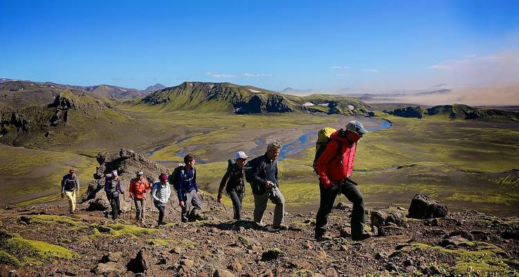 Fila de excursionistas ascienden por una pendiente rocosa que domina un valle volcánico verde y un río sinuoso