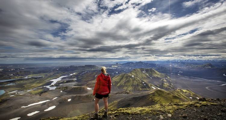 Un senderista solitario con chaqueta roja mira hacia el horizonte desde una cumbre rocosa contemplando picos volcánicos sin fin y lagos