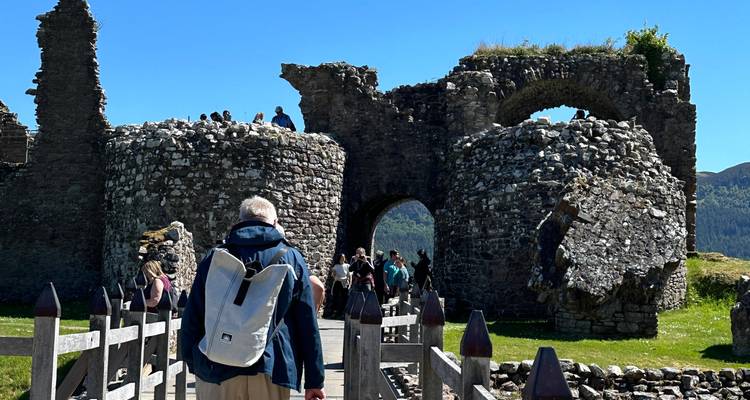 Visitors crossing a wooden bridge toward the stone ruins of Urquhart Castle on a bright day.