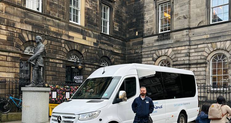 Chauffeur in kilt staand naast een witte toerminibus buiten een historisch stenen gebouw.