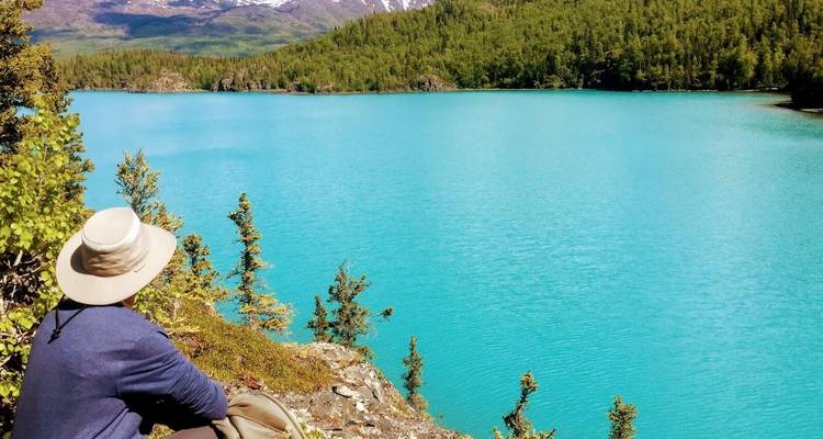 Person sitting by a turquoise lake with mountain views.