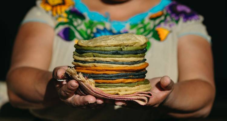 Une femme présente une pile colorée de tortillas artisanales bleues, vertes et jaunes sur ses paumes ouvertes.