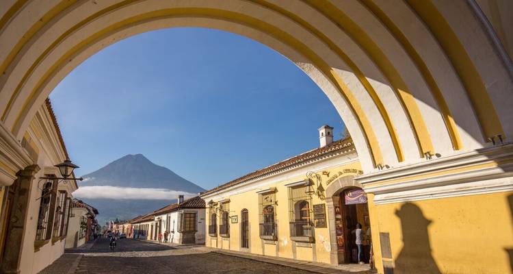L'arche jaune de Santa Catalina encadre une rue pavée avec le lointain Volcán de Agua qui s'élève sous un ciel bleu dégagé.
