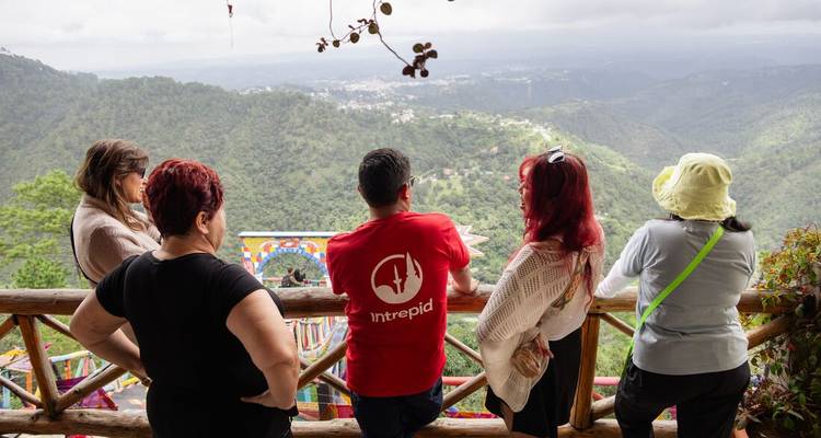 Des voyageurs se tiennent sur un belvédère en bois décoré de mosaïques colorées, contemplant une vallée montagneuse luxuriante.