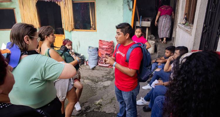 Un guide en chemise rouge parle à un petit groupe dans une modeste cour guatémaltèque avec des sacs de charbon empilés à proximité.