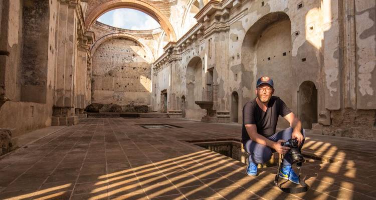 Fotógrafo se agacha con su cámara dentro de las ruinas iluminadas por el sol de una gran iglesia colonial, sombras con patrones en el suelo de piedra.