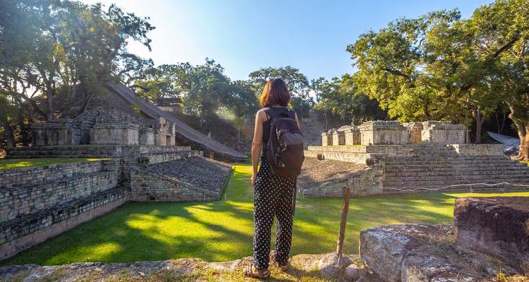 Traveller with backpack looks over expansive ancient stone structures of Copán Ruins bathed in morning light.
