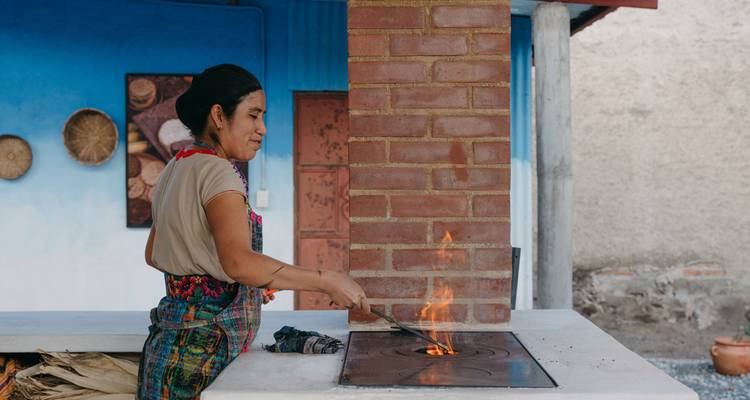 Local woman cooks over an open flame on a traditional wood stove set against a painted blue wall.