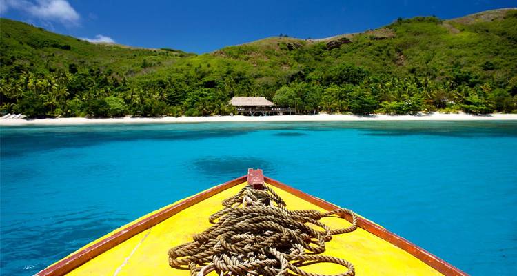 View from a boat's yellow bow across crystal-clear turquoise lagoon toward a lush island shoreline.
