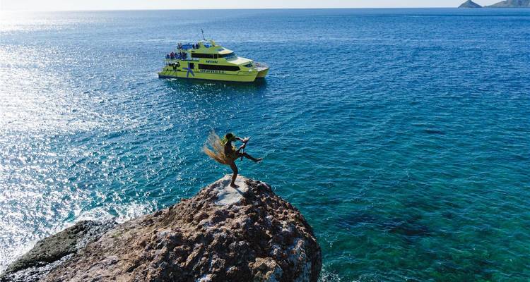 Woman in grass skirt dances on a rocky outcrop high above turquoise sea with tour boat below.