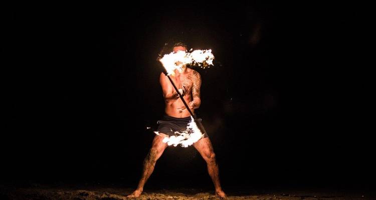 Un artiste torse nu fait tournoyer un bâton enflammé sur une plage tranquille, le feu illuminant la nuit obscure.