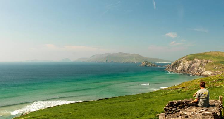 Un viajero se sienta en un muro de piedra con vistas al mar turquesa vibrante y la costa irlandesa ondulante.