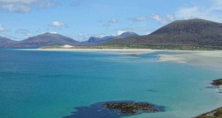 Baie turquoise et vaste plage de sable blanc encadrées par des collines ondulantes et des pics lointains par temps clair.