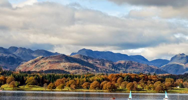 Serene meer met herfstgekleurde bomen en verre Lake District bergen weerspiegeld onder dramatische wolken.