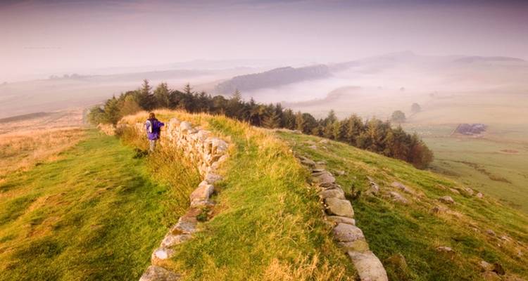Persona caminando junto a un muro de piedra en un paisaje montañoso.