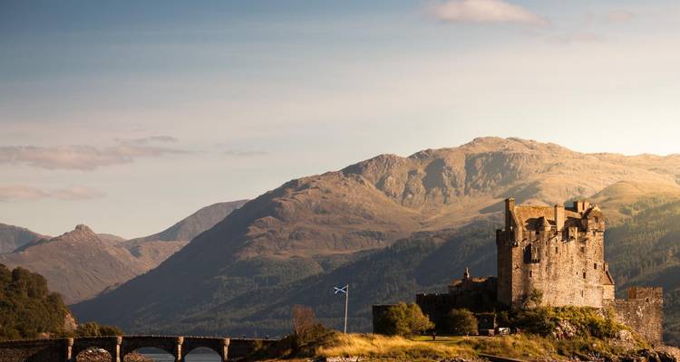 Castillo de piedra con bandera escocesa que domina un puente y un paisaje montañoso bañado por luz cálida.